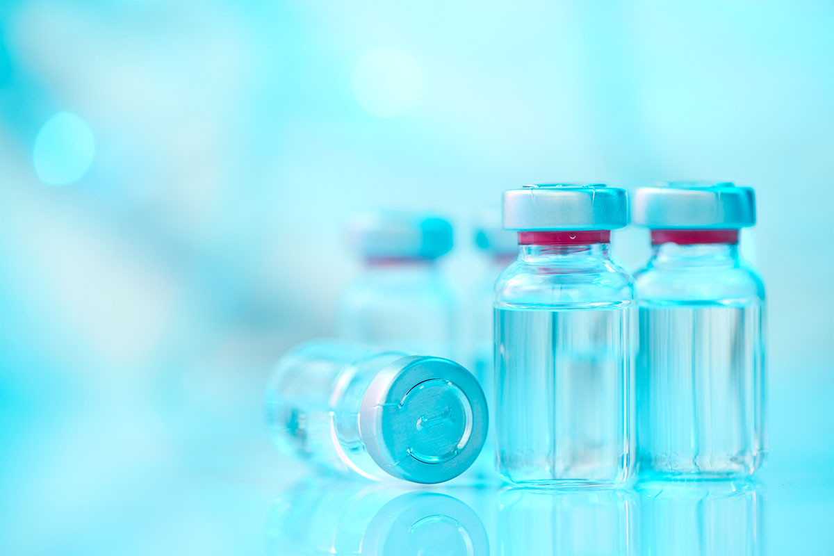 Glass vaccine vials with silver caps and red seals on a reflective surface against a soft blue background, one vial lying on its side