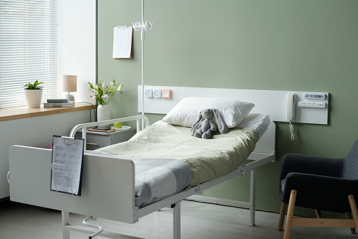An empty adjustable bed in a long-term care room with sage green walls, an IV pole, a chart clipboard on the footboard and an armchair beside it