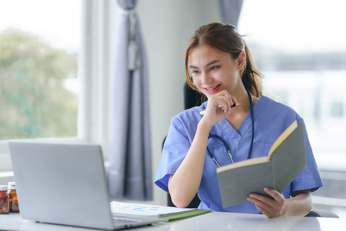 A clinical research nurse in scrubs reviewing patient data on a laptop in a clinic office