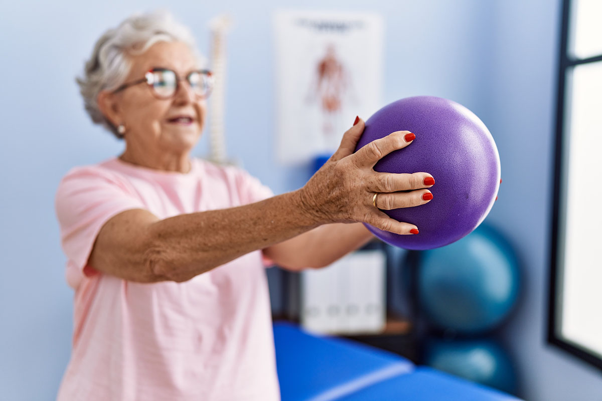 An older woman with grey hair and glasses holding a purple exercise ball at arm's length during a physical therapy session